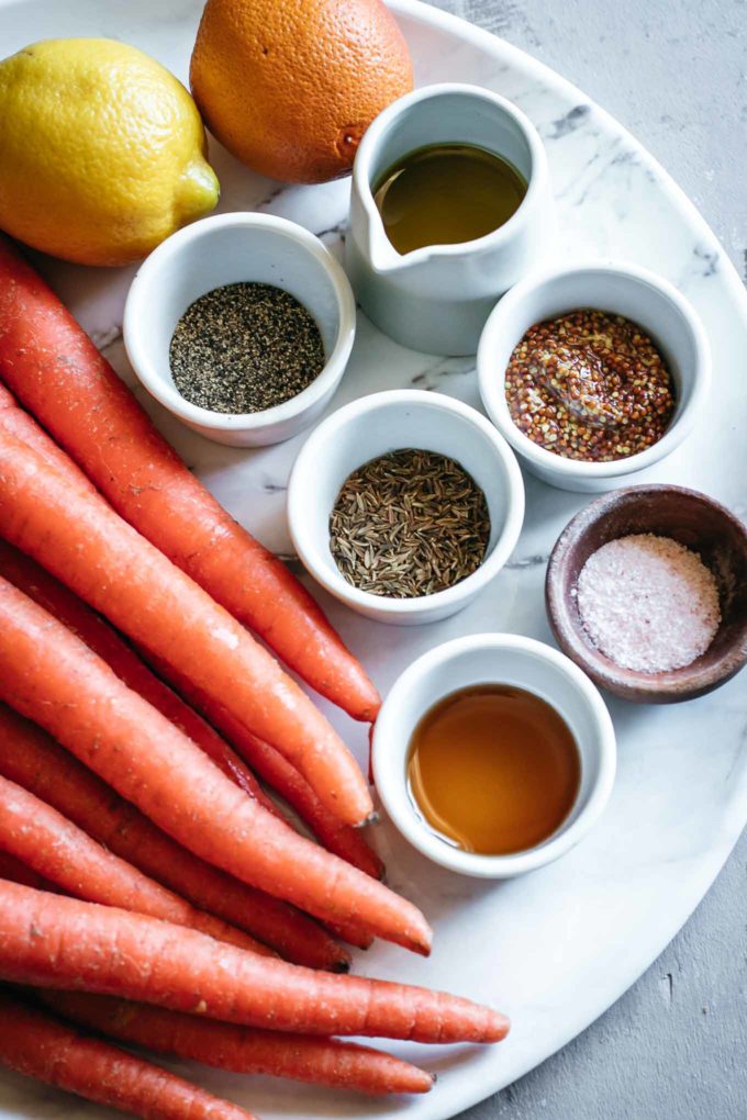bowls of oil, maple syrup, and spices next to pile of carrots, lemon, and an orange