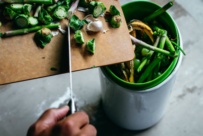 a hand throwing vegetable food scraps into a white compost pail