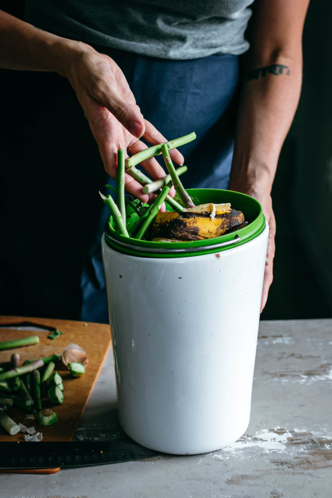 a hand pouring food scraps into a compost bin on a wood table