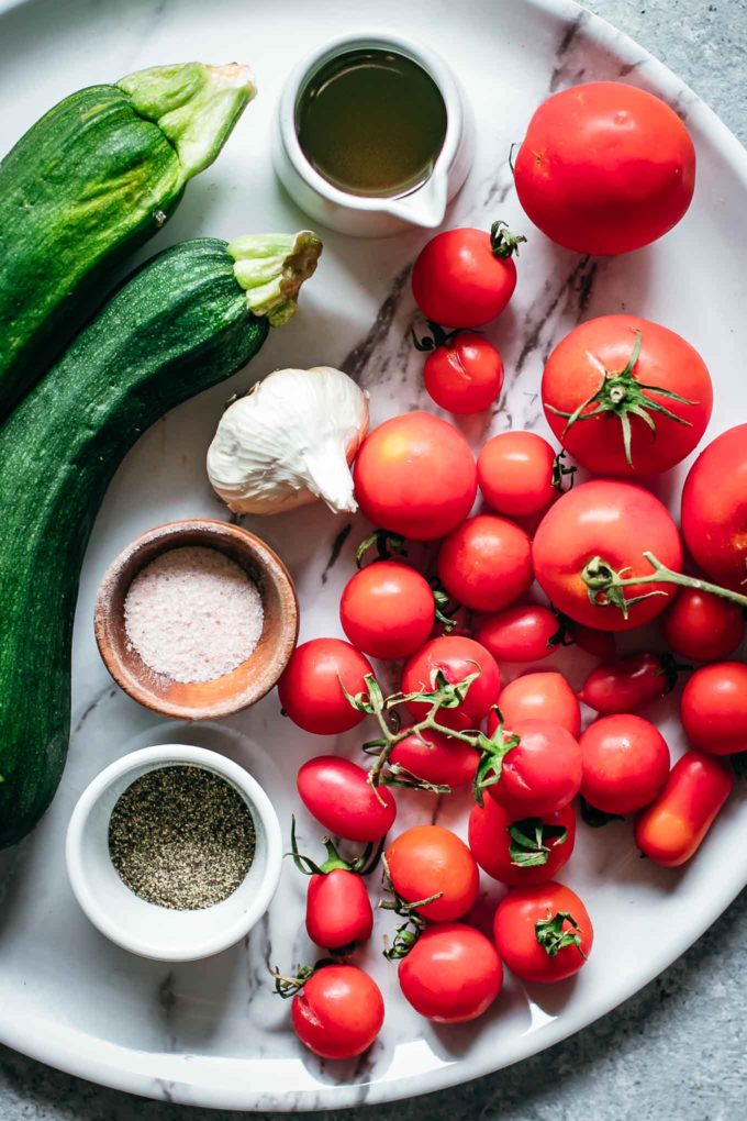 tomatoes, zucchini, garlic, herbs, oil, salt, and pepper on a white countertop