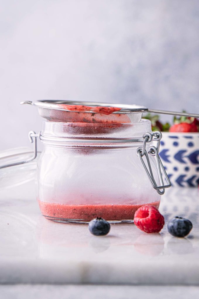 a mesh strainer with strawberry puree over a glass bowl on a white table