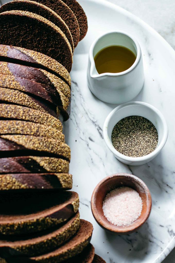 sliced loaf of pumpernickel bread, olive oil, salt and pepper on a white table