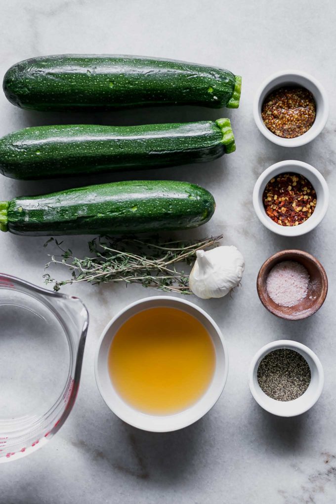 whole zucchini and bowls of water, vinegar, salt, pepper, mustard, garlic, and fresh herbs on a white table