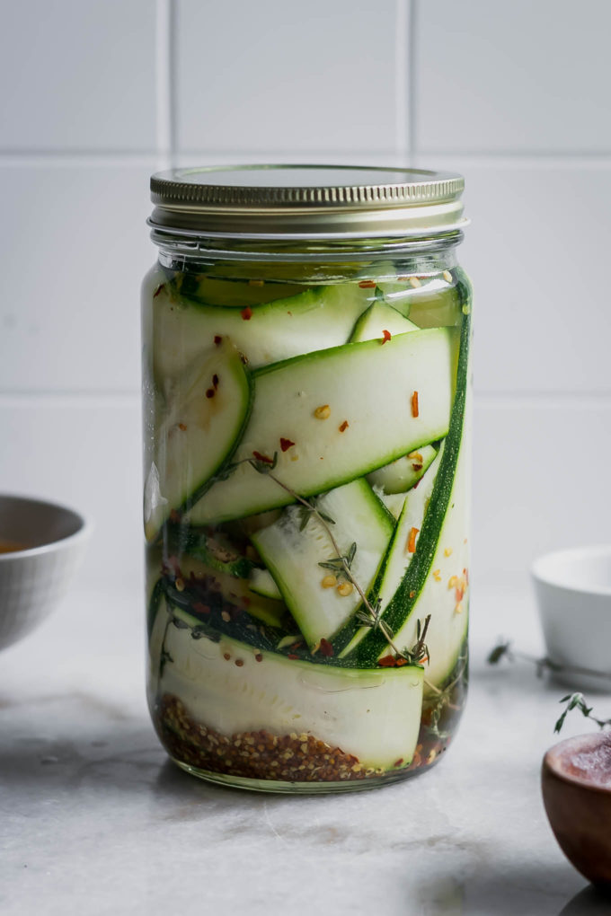 pickled thinly sliced zucchini in a glass jar with pickling brine on a white counterop