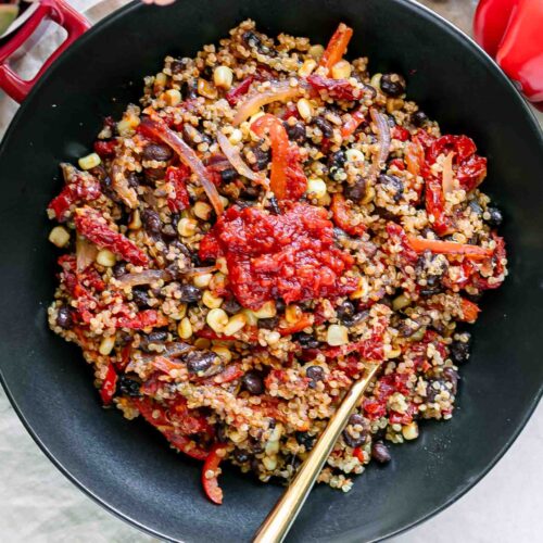a quinoa bowl with black beans, bell peppers, onion, sun-dried tomatoes, and spicy harissa sauce on a white table with a gold fork