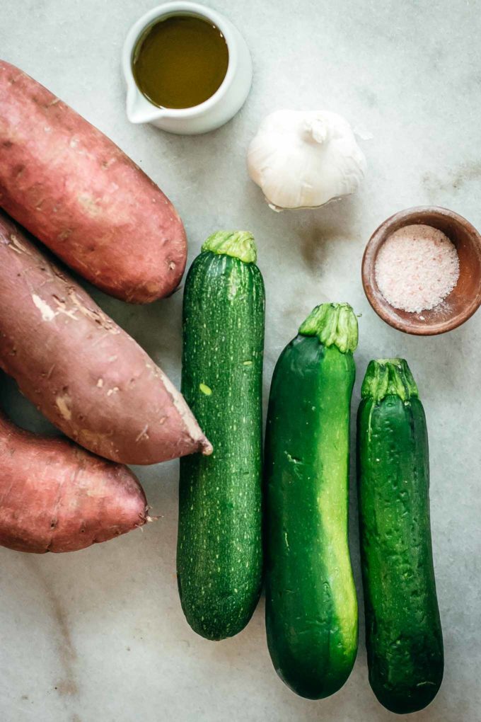 sweet potatoes, zucchini, garlic, and bowls of garlic and olive oil on a white countertop