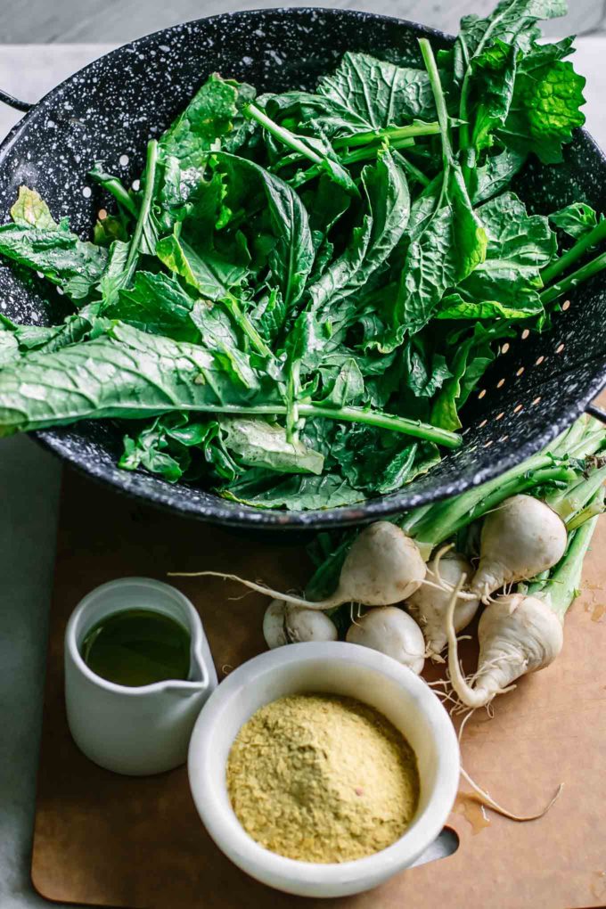 turnip greens in a colander next to turnips, olive oil and nutritional yeast