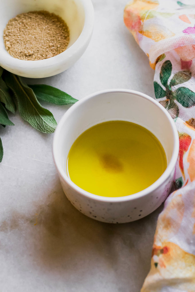 a bowl of butter and sage sauce on a white table with a floral napkin and fresh sage leaves