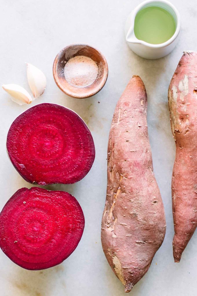 a sweet potato, cut beet, garlic cloves, and bowls of oil and salt on a white table