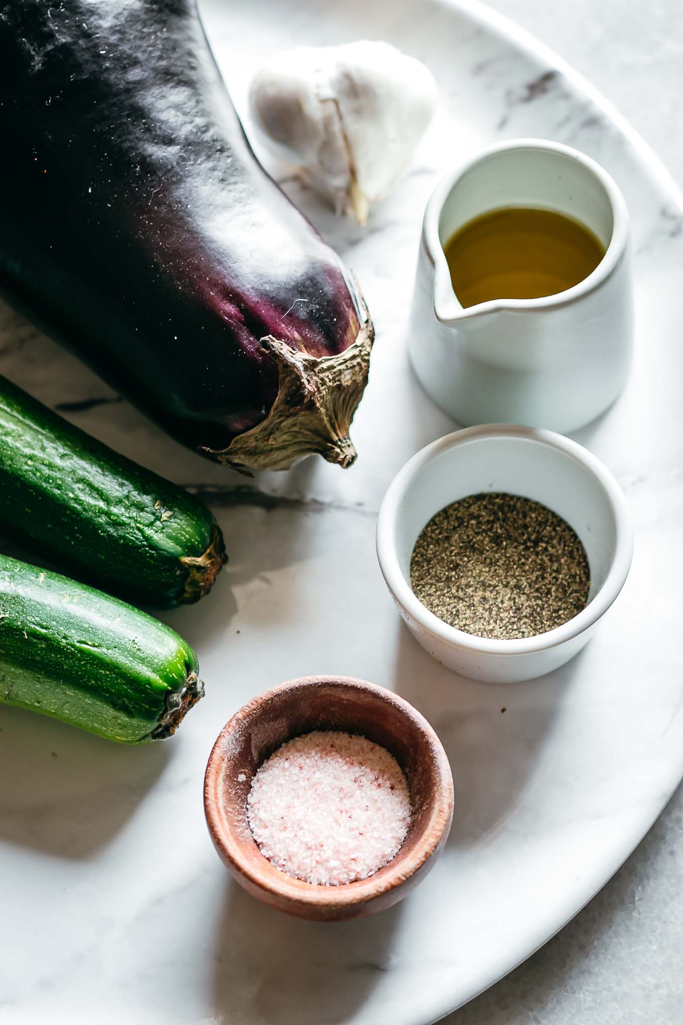 eggplant, zucchini, garlic, and bowls of oil, salt, and pepper on a white table