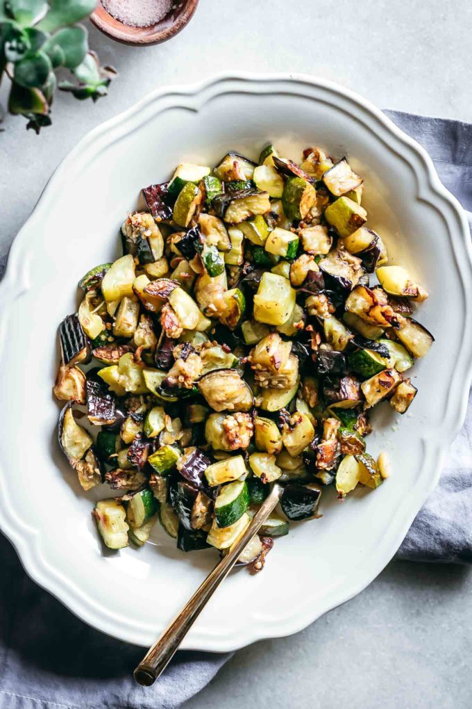roasted eggplant and zucchini cubes on a white plate on a white table with a gold fork