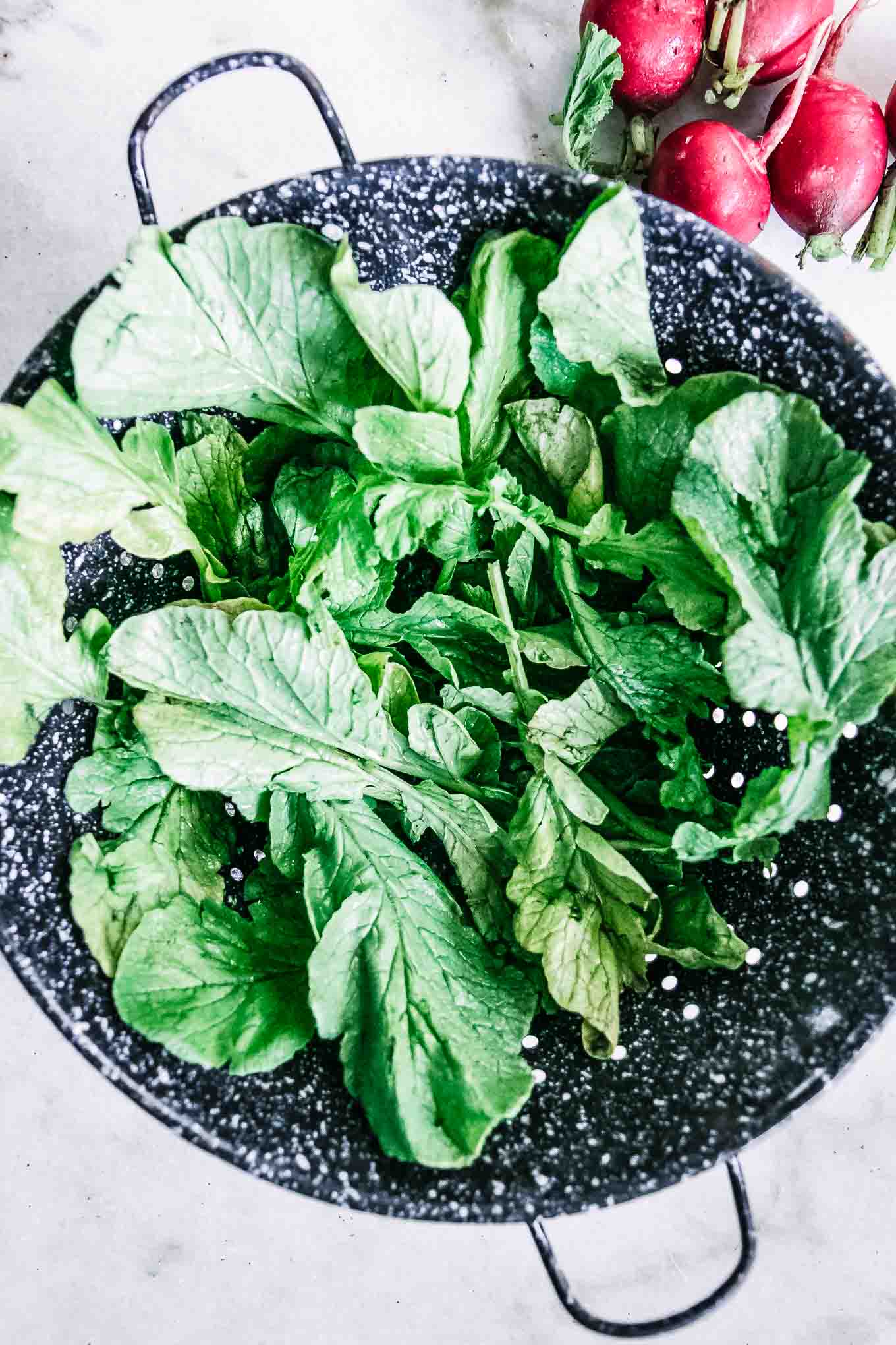 washed radish leaves drying in a blue colander on a white table with a bunch of radishes