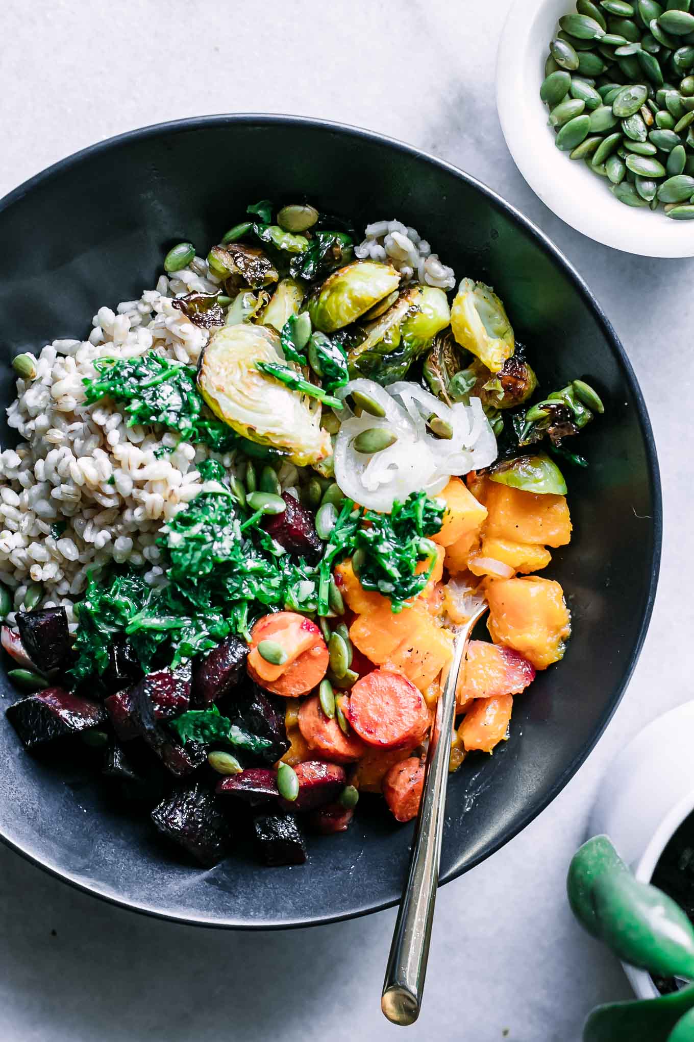 a bowl of roasted vegetable barley with herb sauce, a gold spoon and a bowl of pumpkin seeds