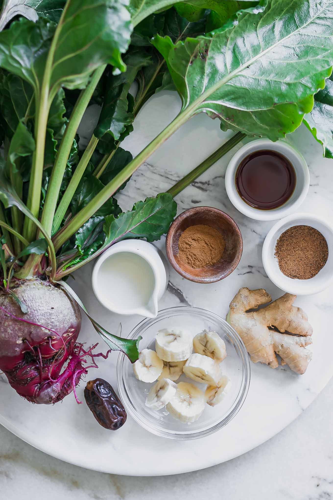 a beet, date, and bowl of milk, banana slices, ginger, cinnamon, nutmeg, and vanilla on a white table