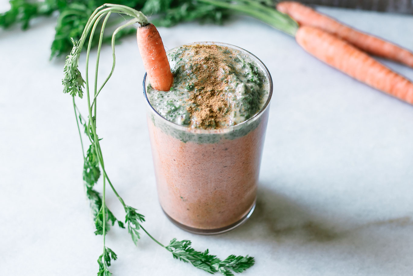 an orange carrot smoothie with a green carrot tops smoothie layer on top in a glass on a white table