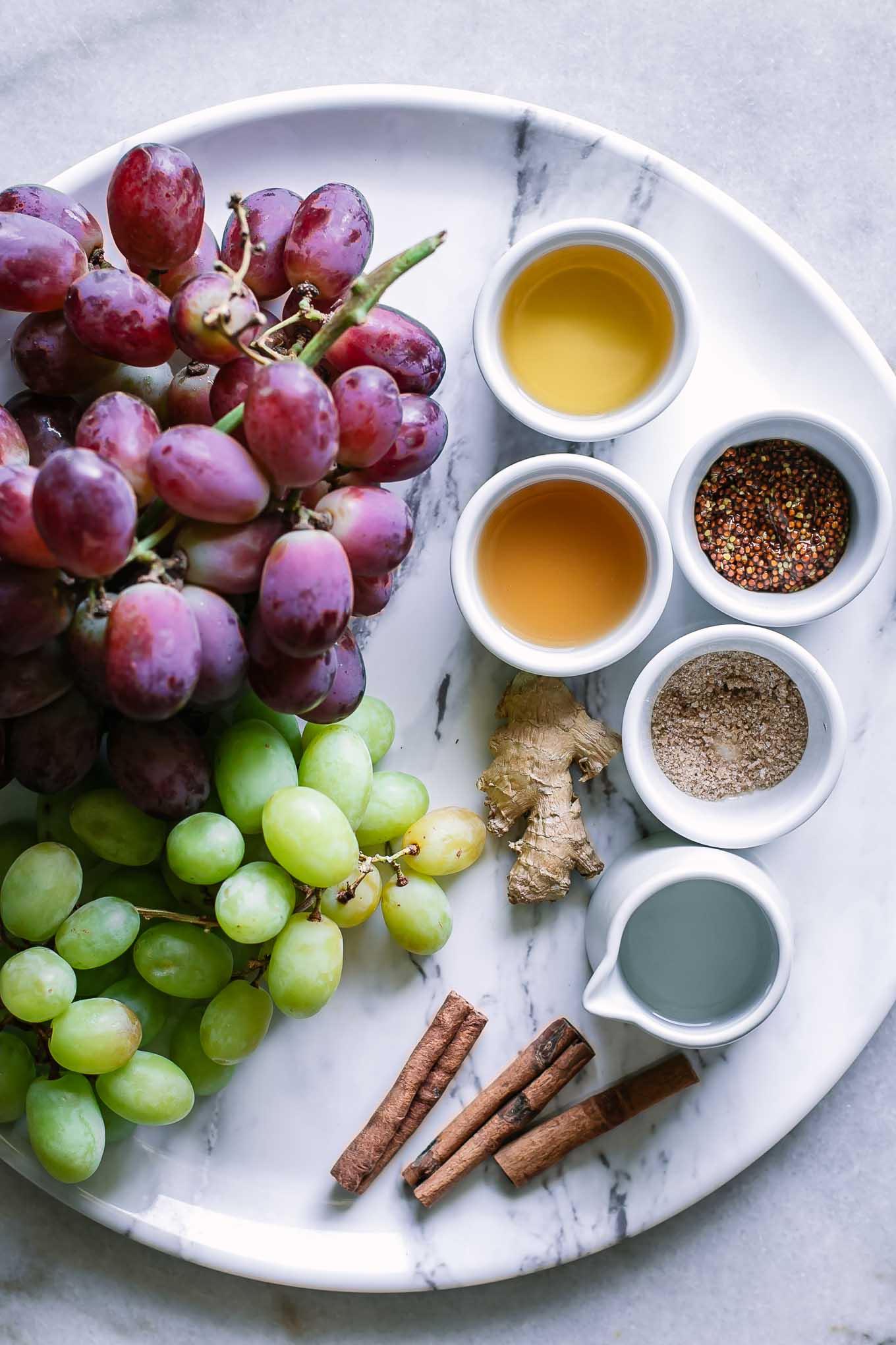 red and green grapes, cinnamon sticks, ginger root, and bowls of water, vinegar, mustard seeds, sugar, and salt on a white table