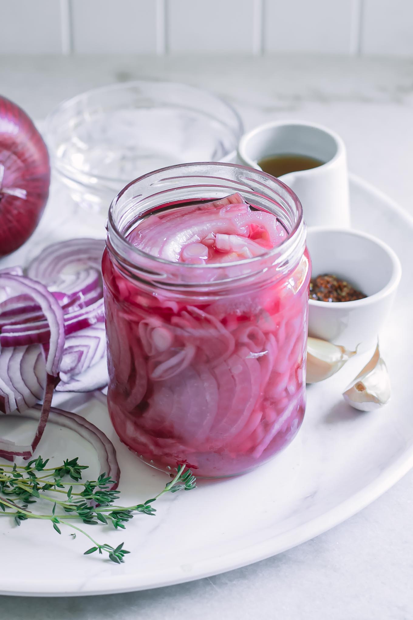 jar of pickled red onions on a plate with onions, oil, garlic, spices and herbs