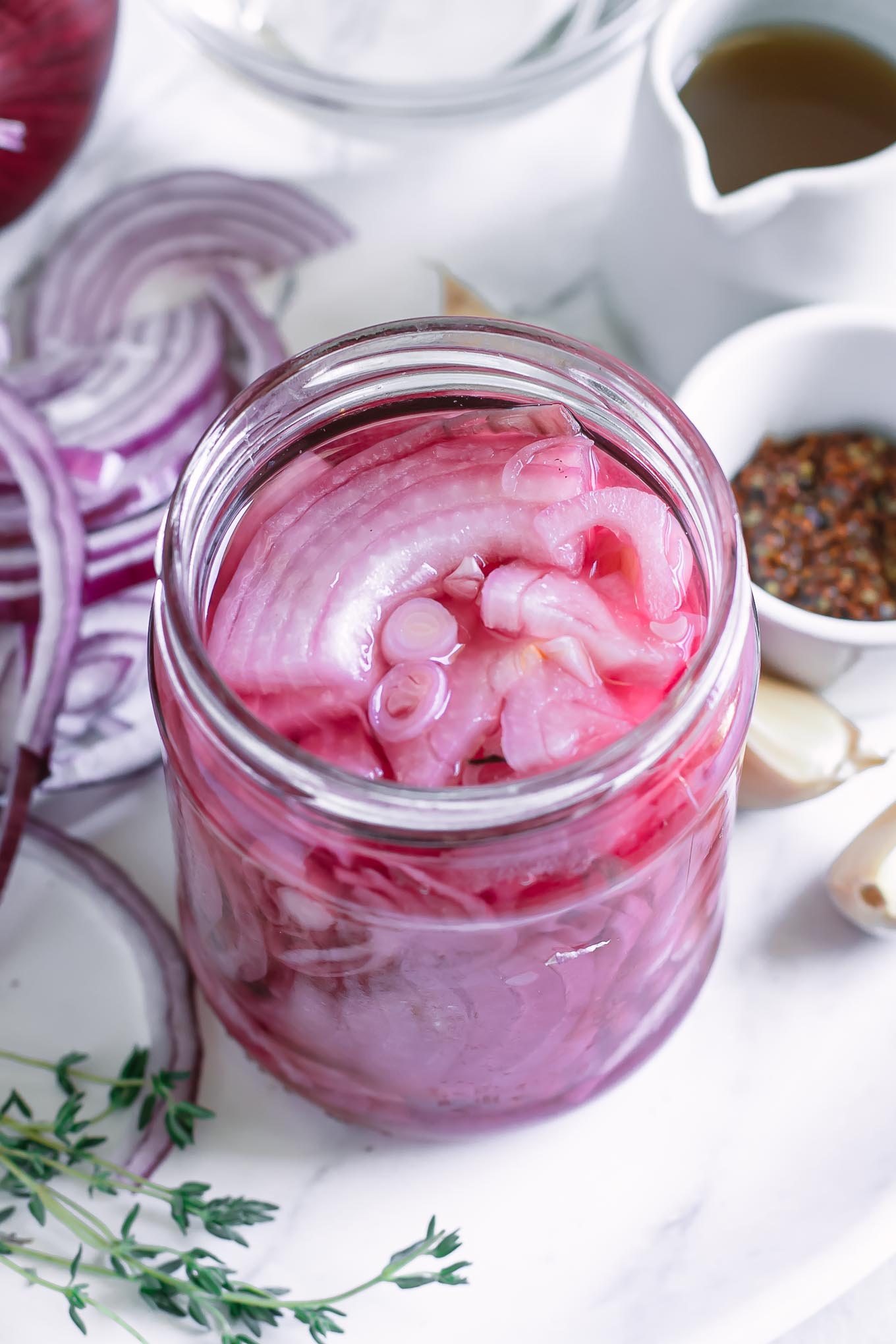 side view of a jar of pickled red onions on a plate with onions, herbs and spices