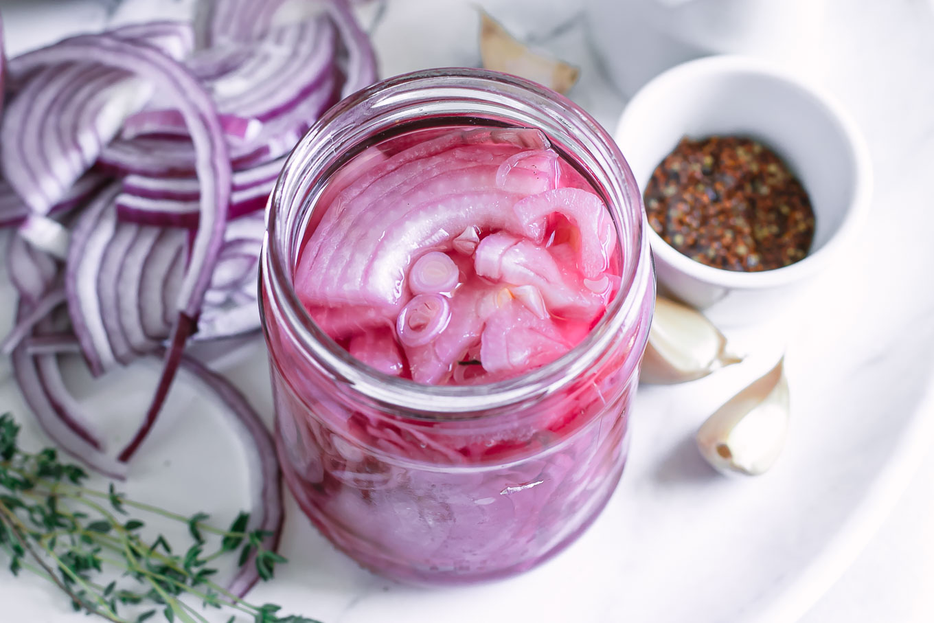 side view of a jar of pickled red onions on a plate with onions, herbs and spices