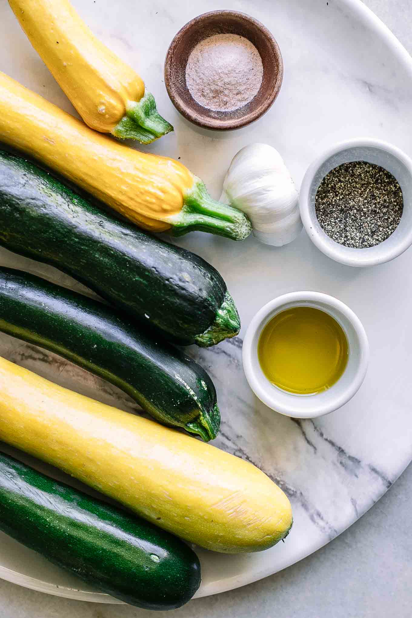 zucchini, summer squash, and bowls of oil, salt, pepper, and garlic on a white table