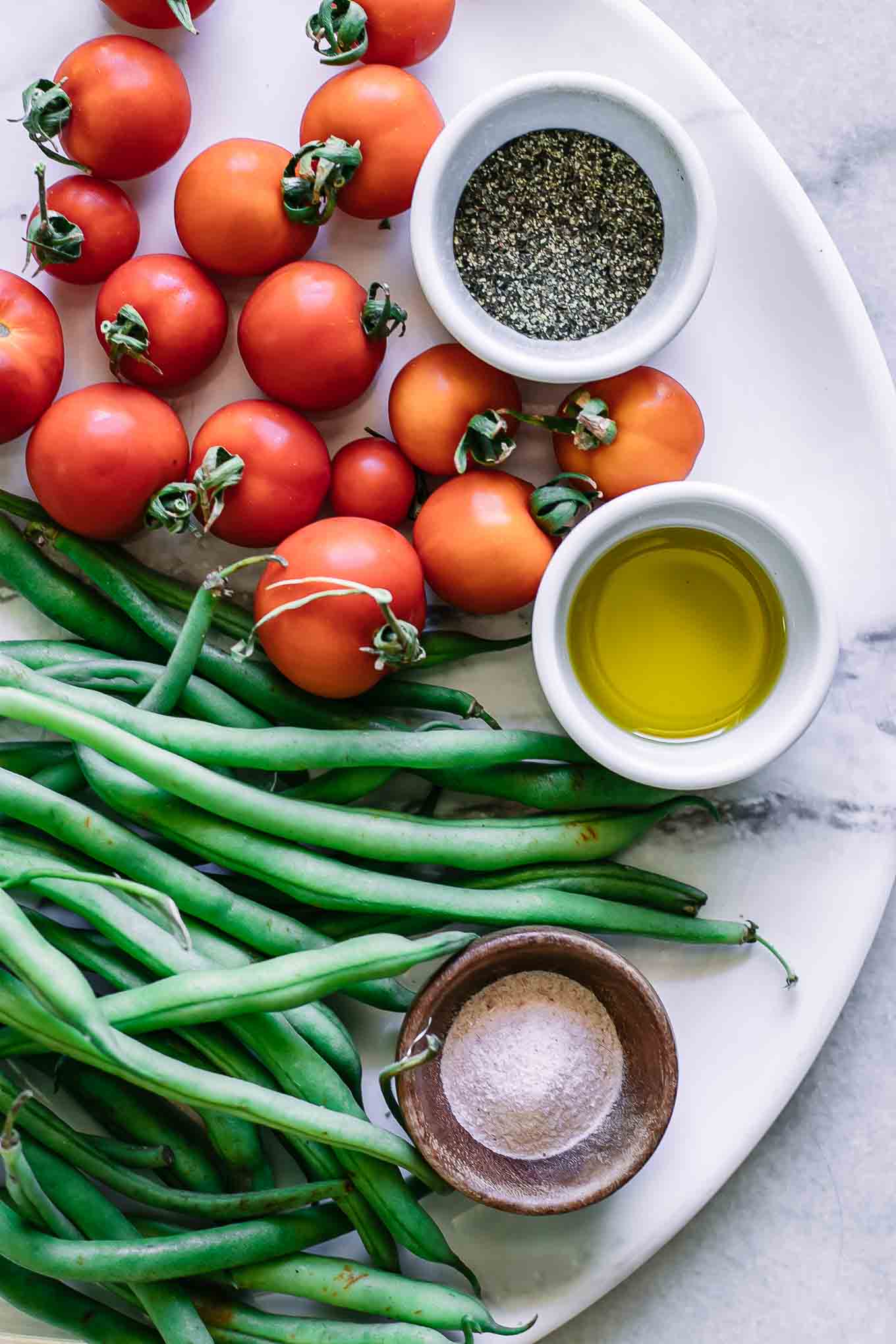 fresh green beans and tomatoes on a white plate with bowls of olive oil, salt, pepper, and garlic