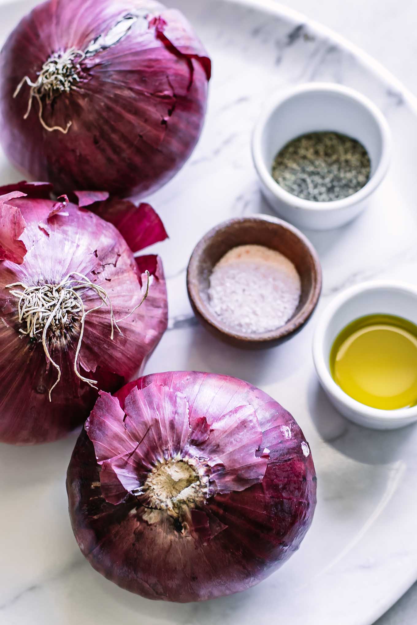 whole red onions on a white plate with bowls of olive oil, salt, and pepper