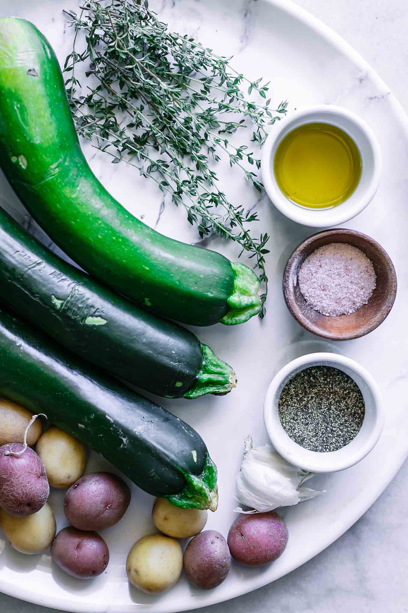 zucchini, small potatoes, fresh herbs, and bowls of oil, salt, and pepper on a white table