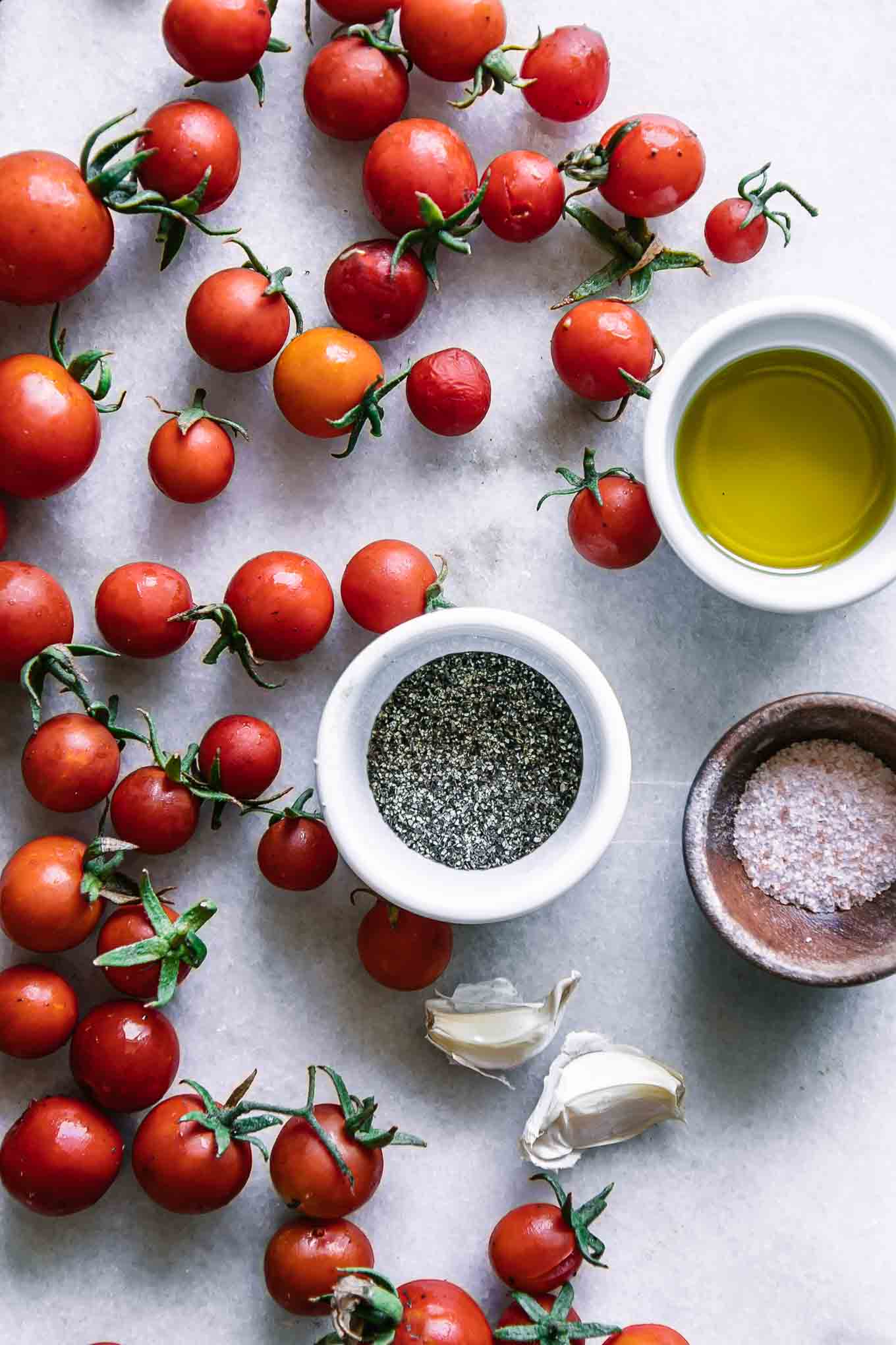 cherry tomatoes on a white table with bowls of oil, garlic, salt, and pepper