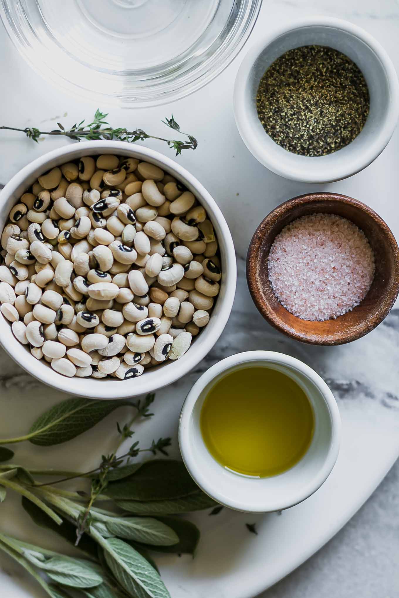 a bowl of dried black eyed peas, fresh herbs, and bowls of oil, salt, and pepper on a white table