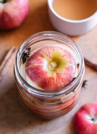 apple slices in to a pickling jar with brine on a wood table with whole apples and bowls of vinegar