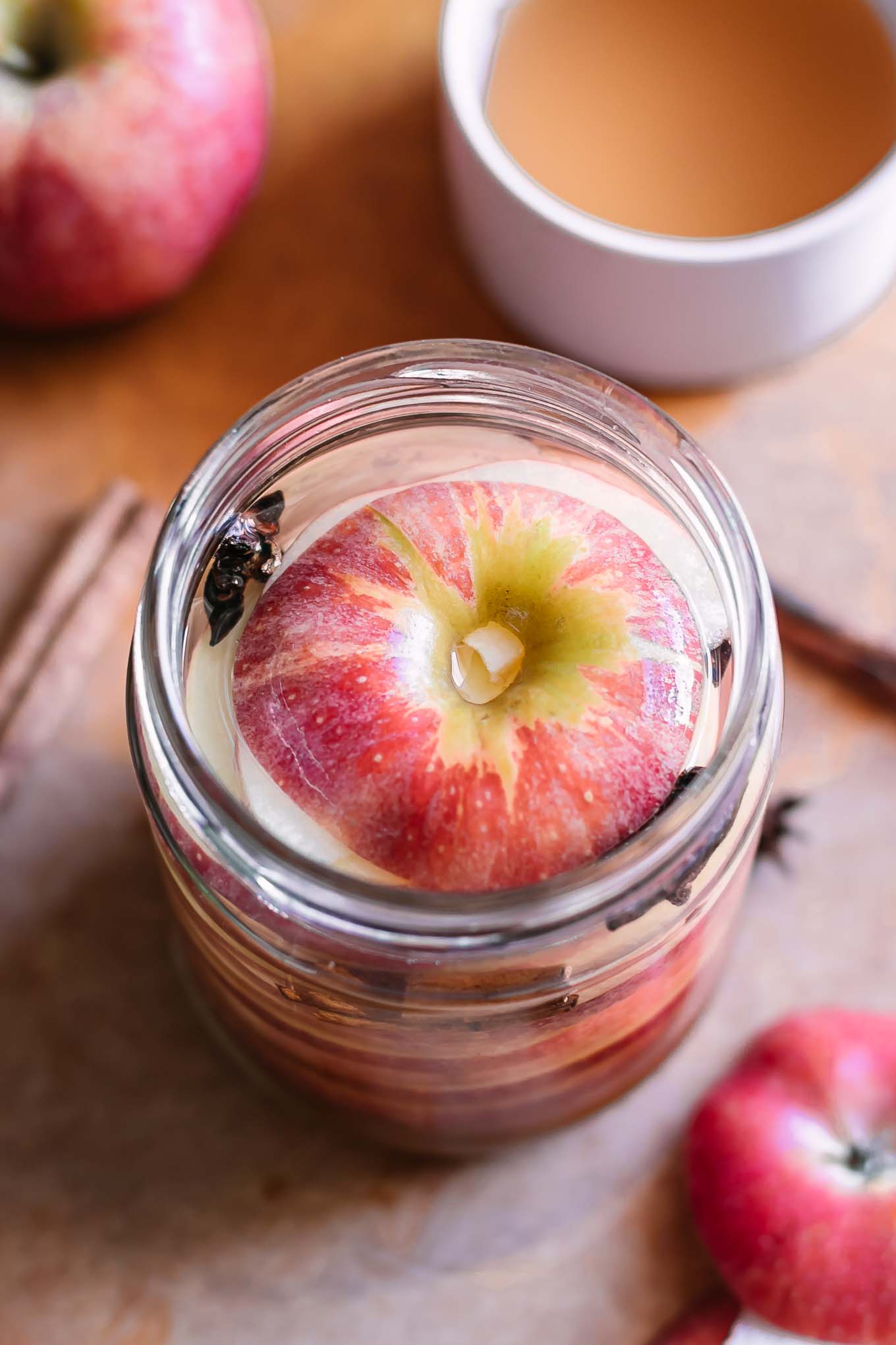 apple slices in to a pickling jar with brine on a wood table with whole apples and bowls of vinegar