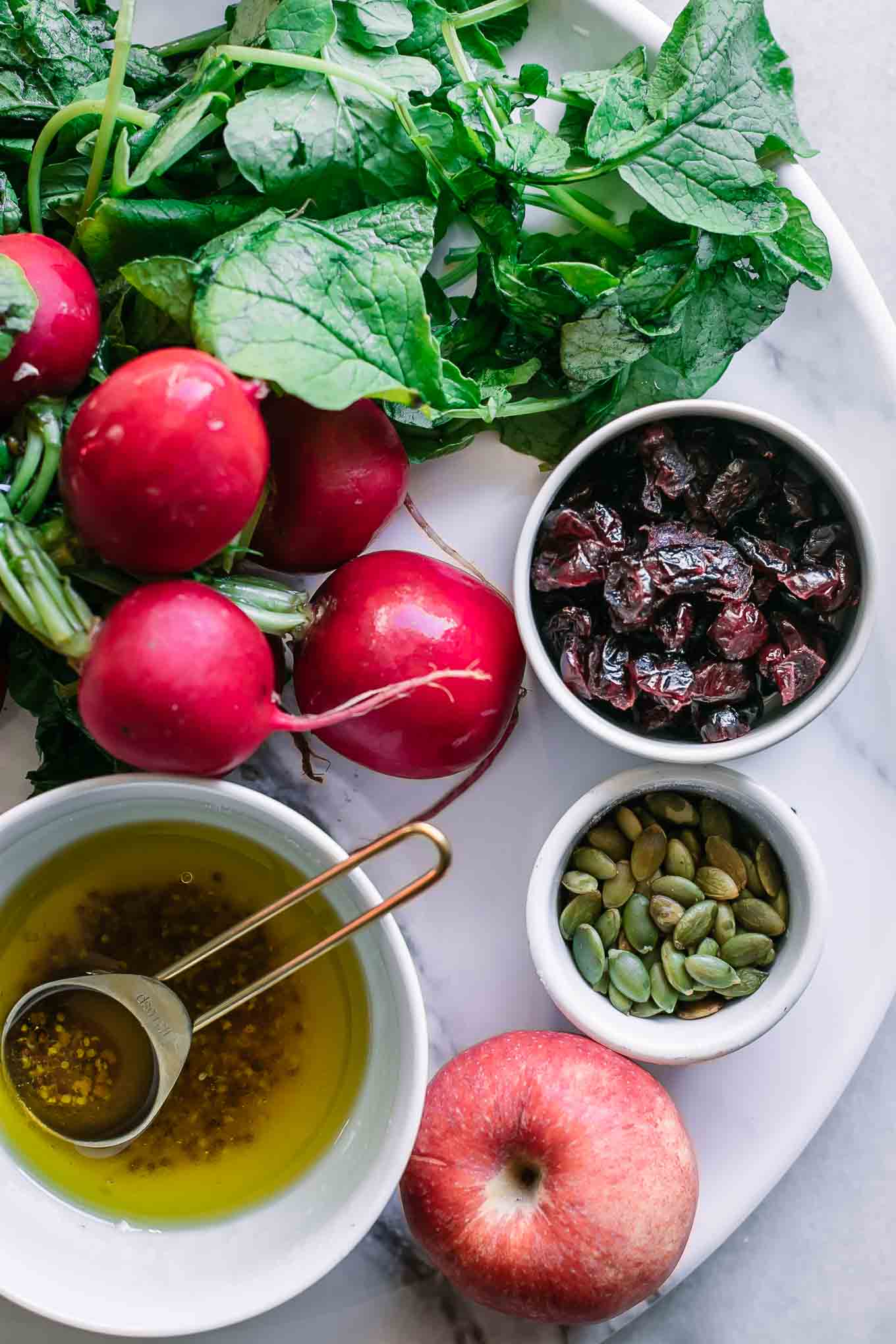 radishes with leaves, an apple, and bowls of cranberries, seeds, and salad dressing on a table
