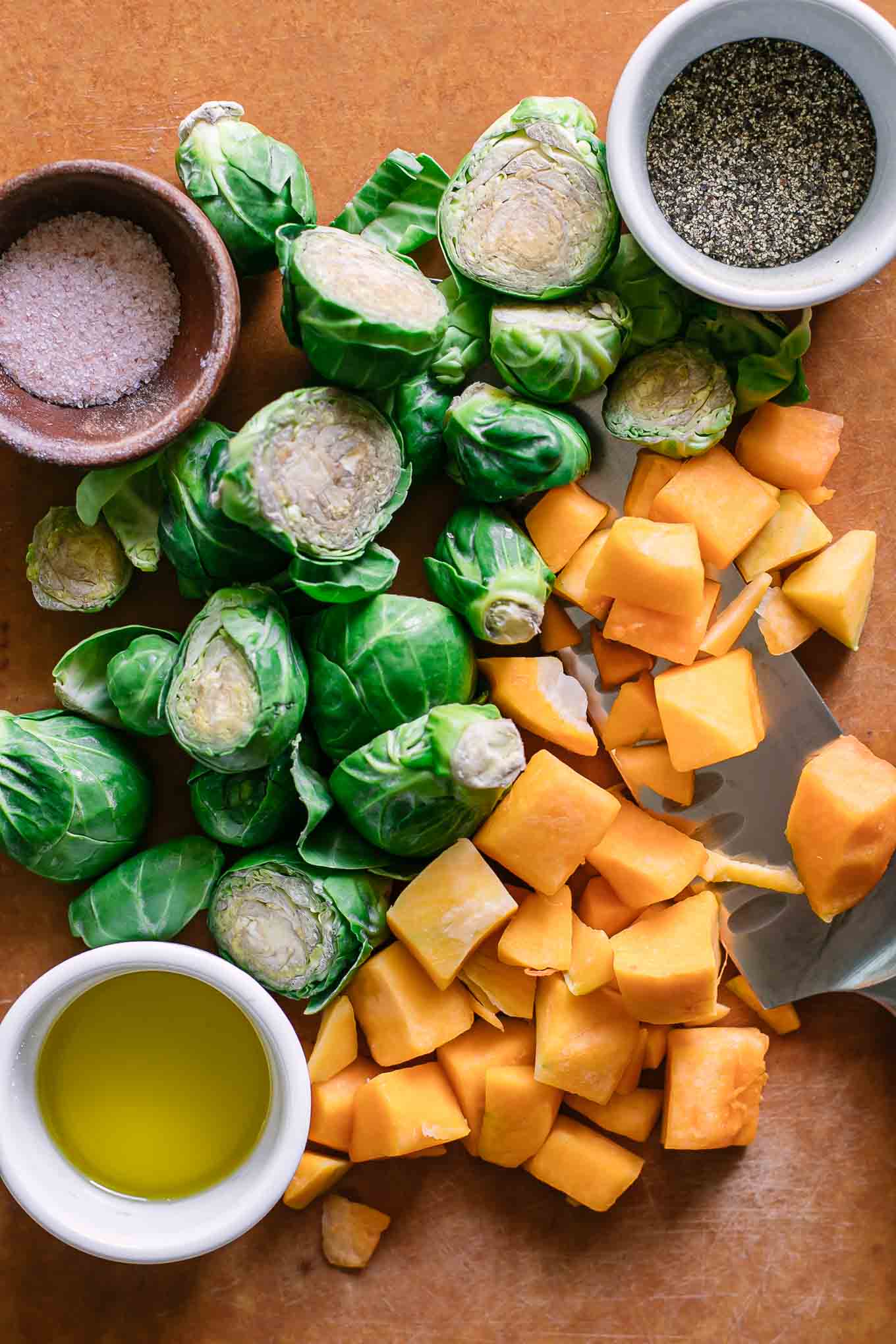 cut brussels sprouts, butternut squash, and bowls of olive oil, salt, and pepper on a cutting board