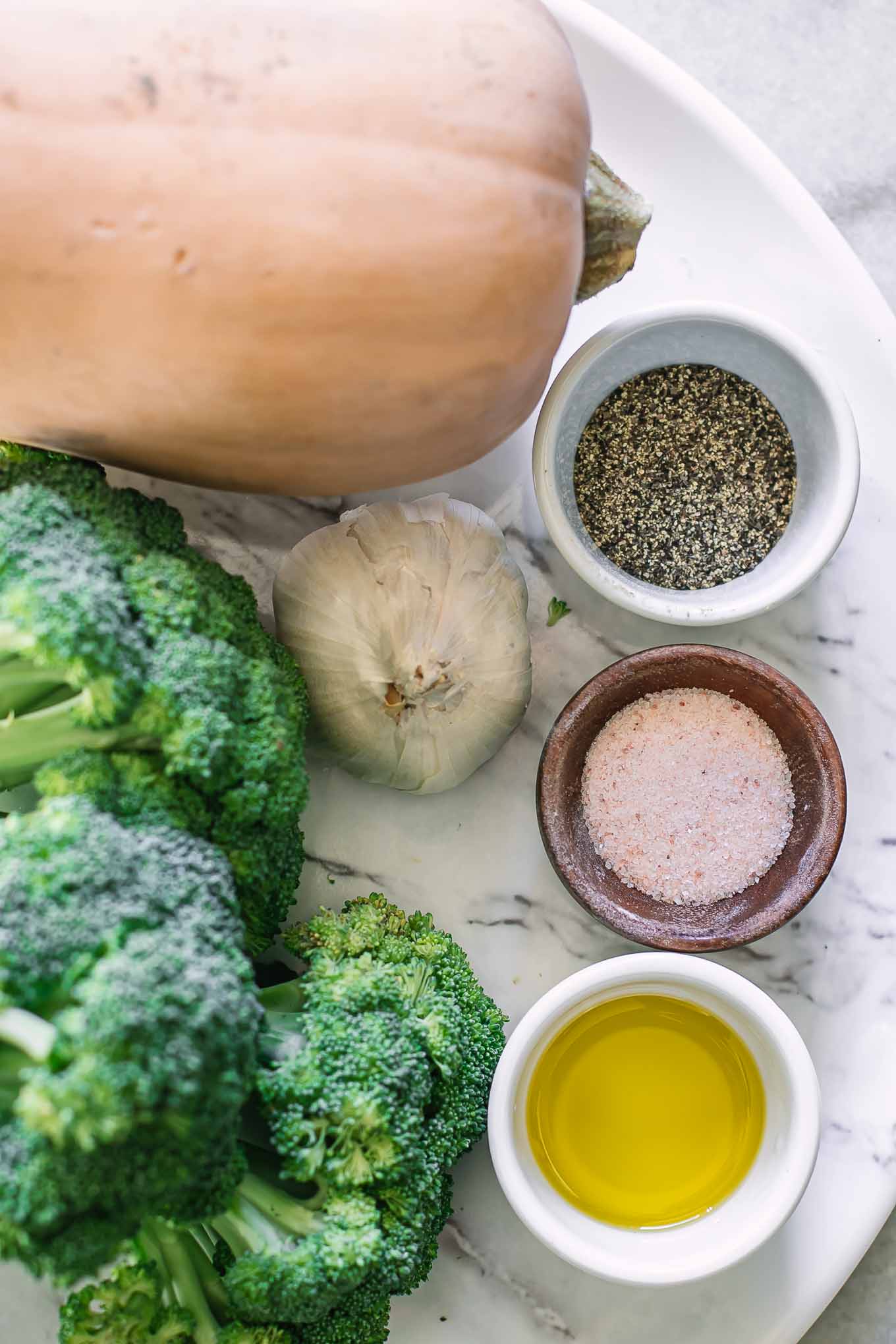 a butternut squash, broccoli stalk, garlic, oil, salt, and pepper on a white table