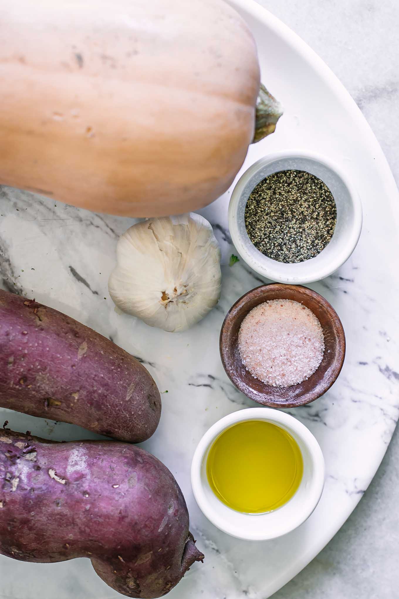 a butternut squash, two sweet potatoes, garlic, and bowls of oil, salt, and pepper on a white table