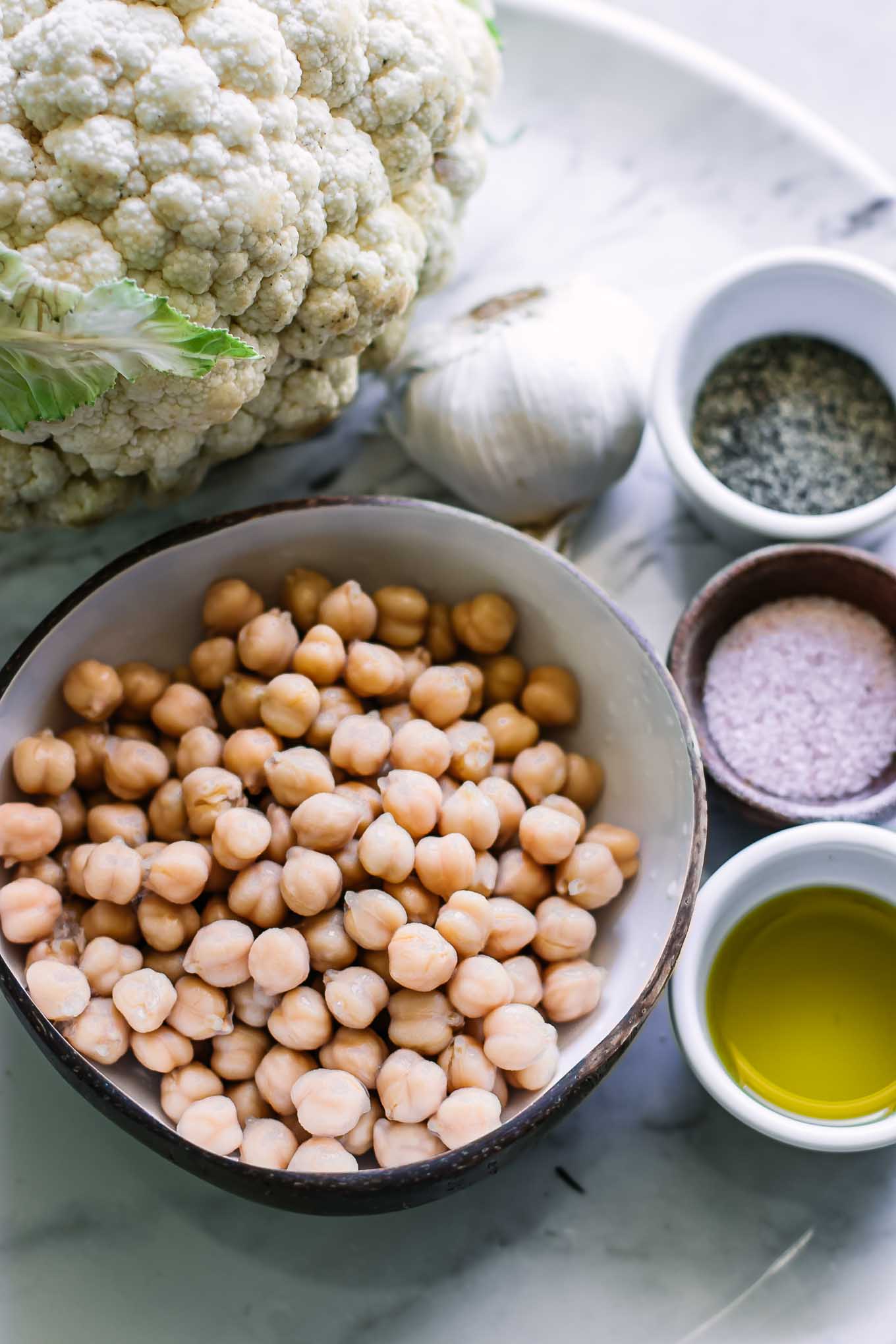 a bowl of chickpeas and a head of cauliflower on a table with oil, salt, and pepper