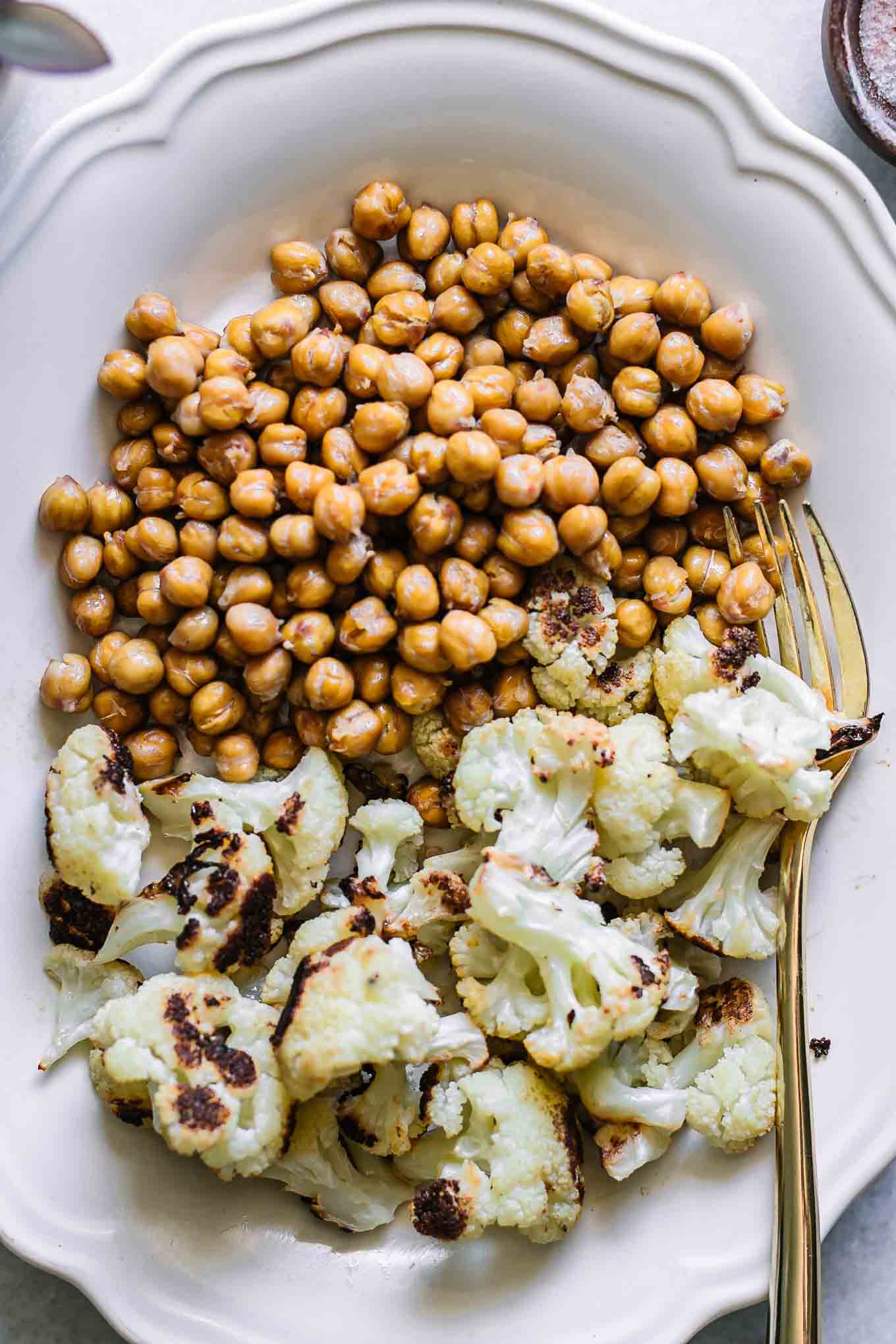 a close up photo of baked cauliflower and chickpeas on a white plate with a gold fork