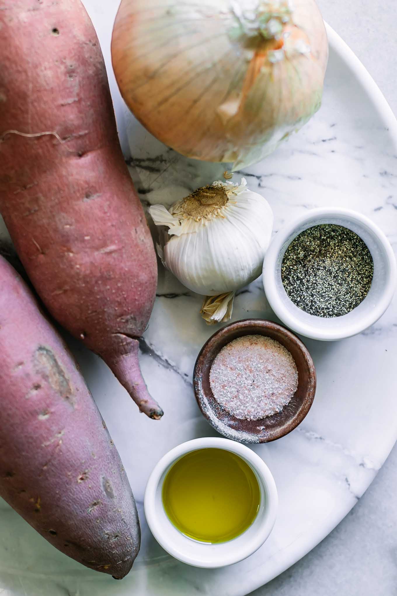 sweet potatoes, an onion, garlic, and bowls of oil, salt, and pepper on a white table