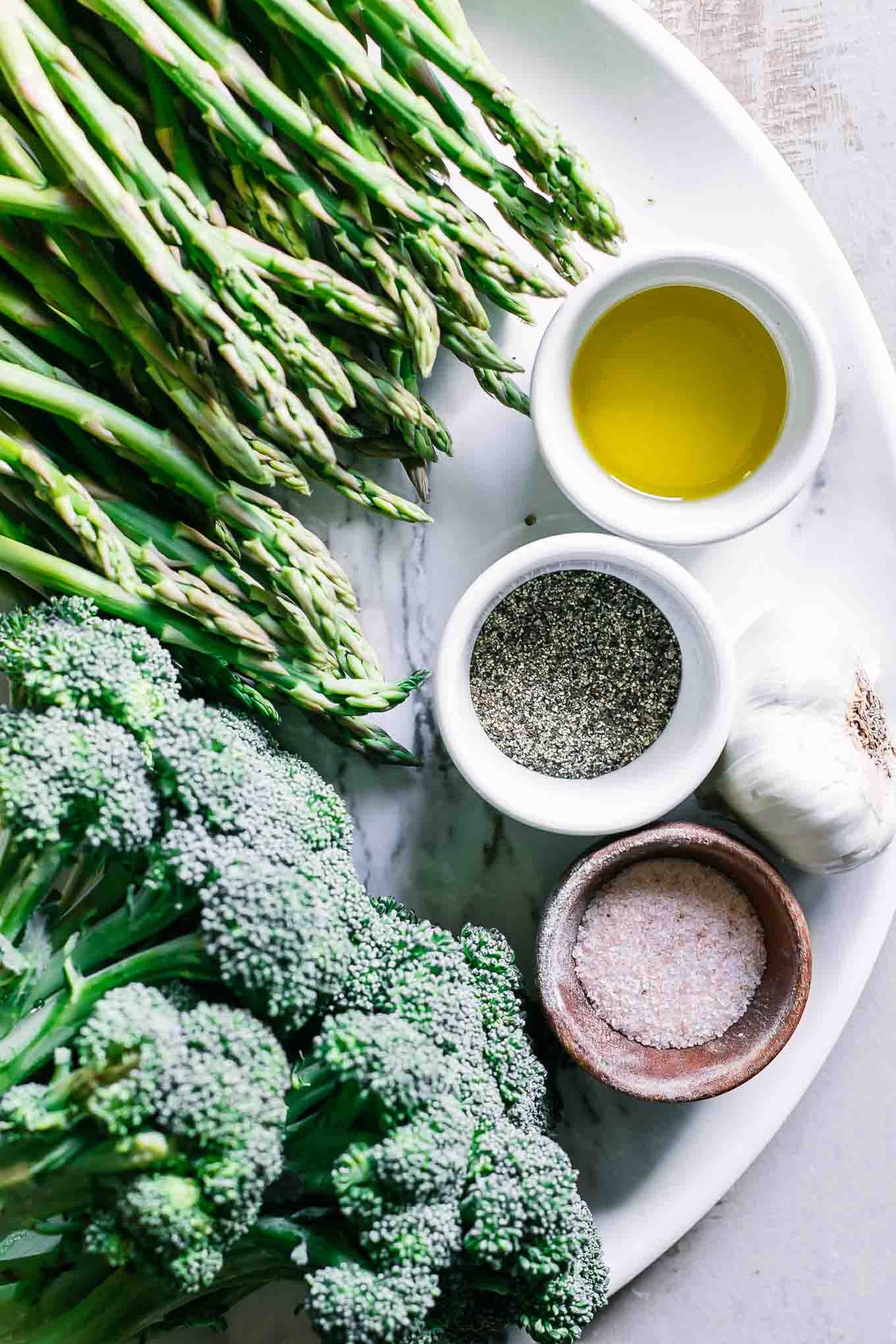 asparagus spears, a head of broccoli, garlic, oil, salt, and pepper on a white table