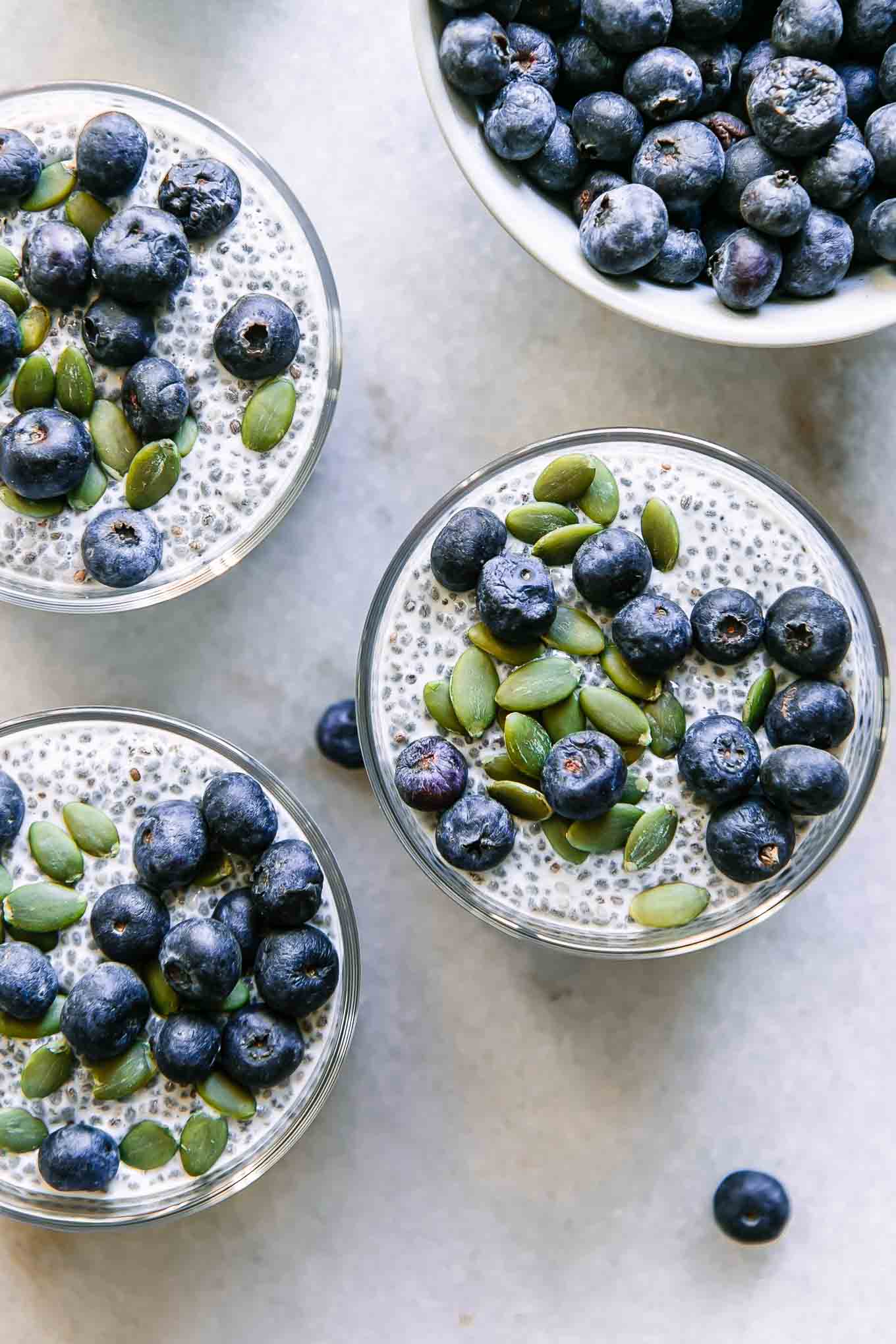 three jars of chia seed pudding with blueberries and pumpkin seeds on a white table