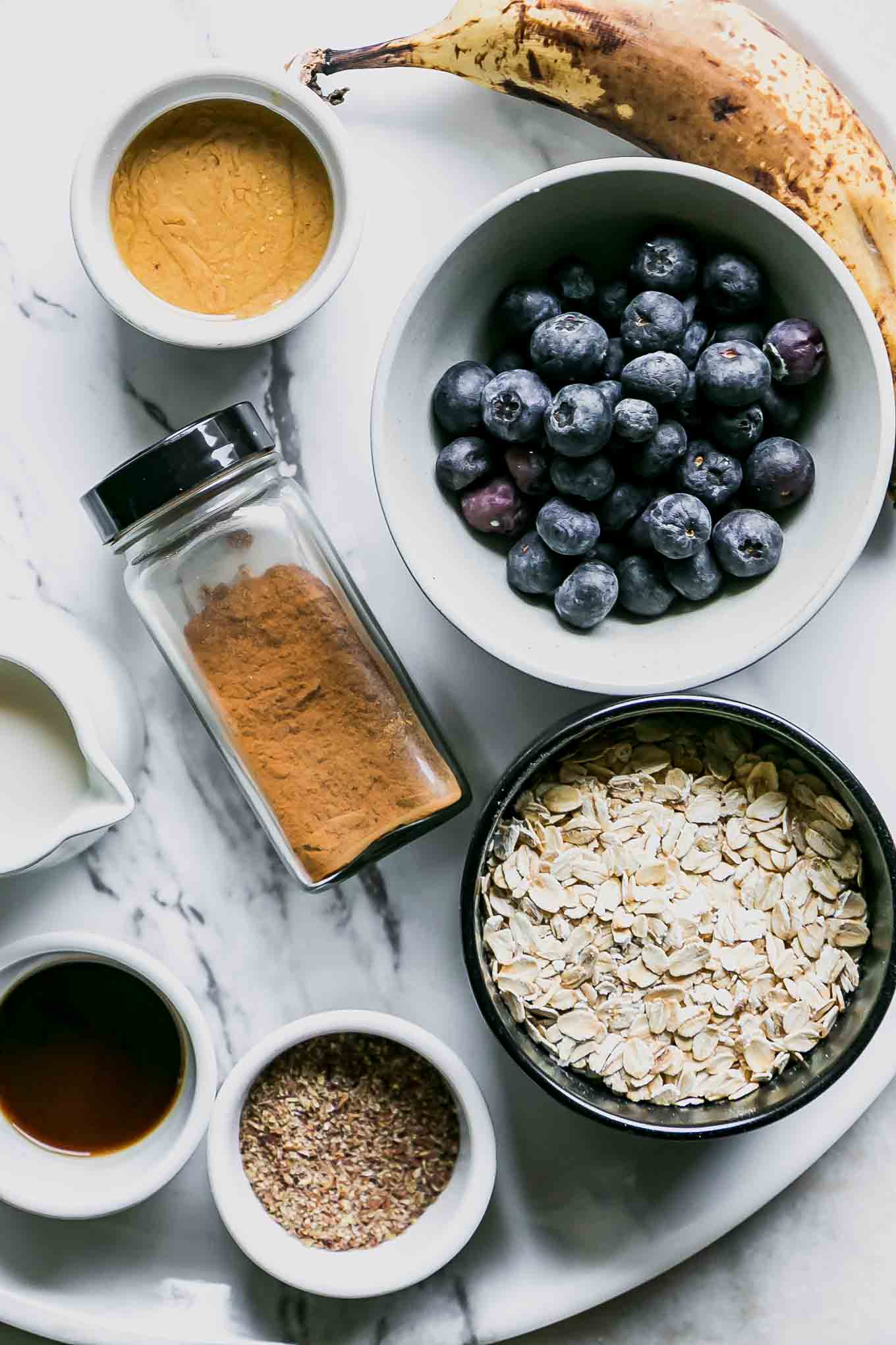 bowls of blueberries, oats, vanilla, flax seeds, milk, and a banana on a white table for a smoothie