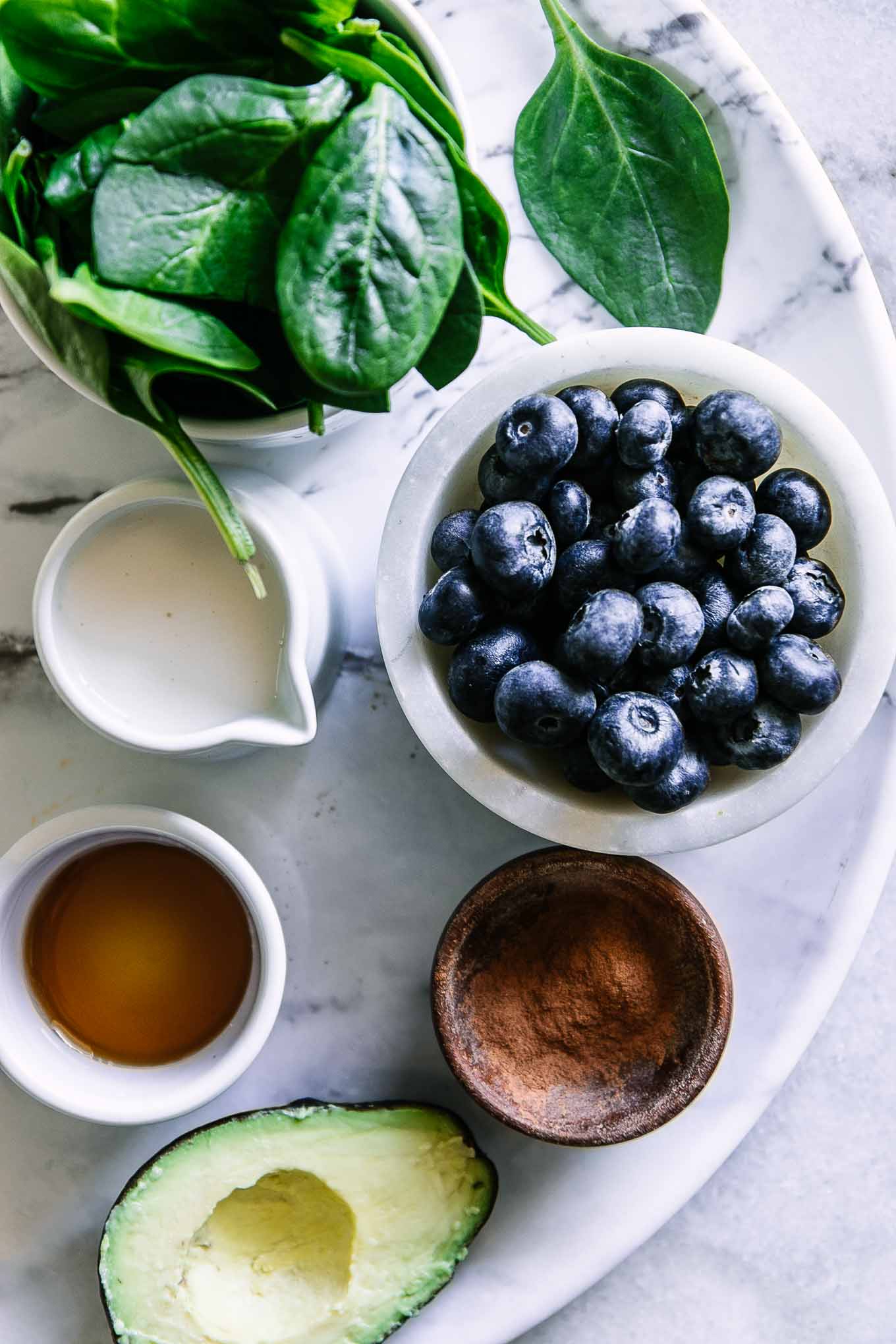 bowls of blueberries, spinach, milk, vanilla, cinnamon, and an avocado on a white table for a smoothie