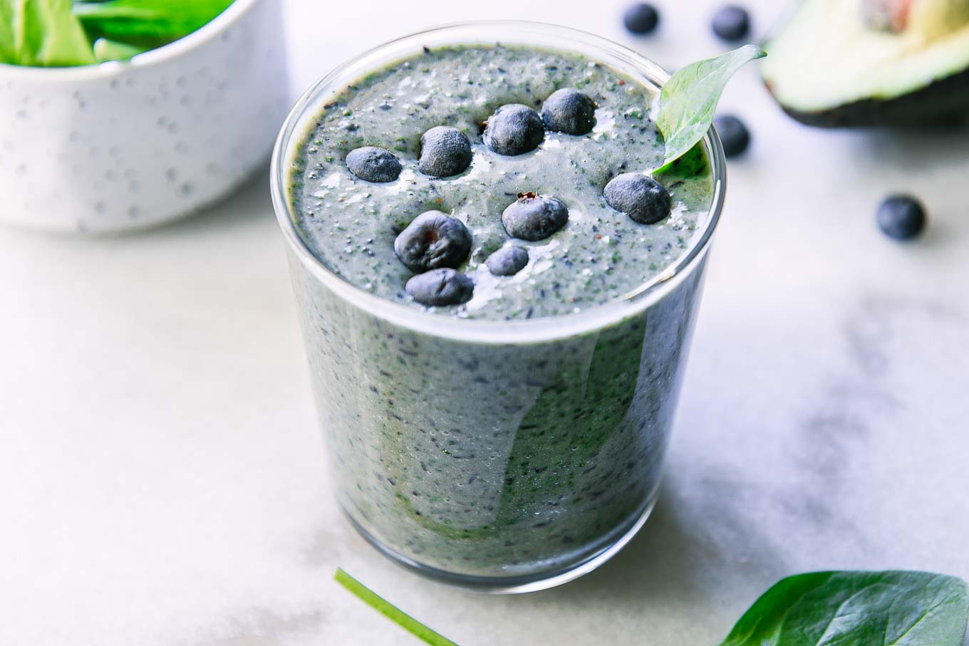 a spinach blueberry smoothie in a glass with blueberries and a spinach leaf as garnish on a white table