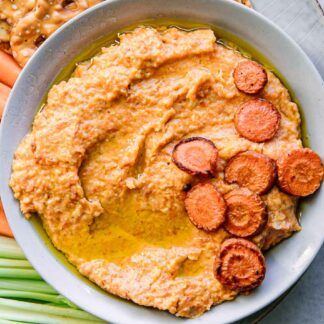 a bowl of carrot hummus dip on a plate with vegetables for dipping