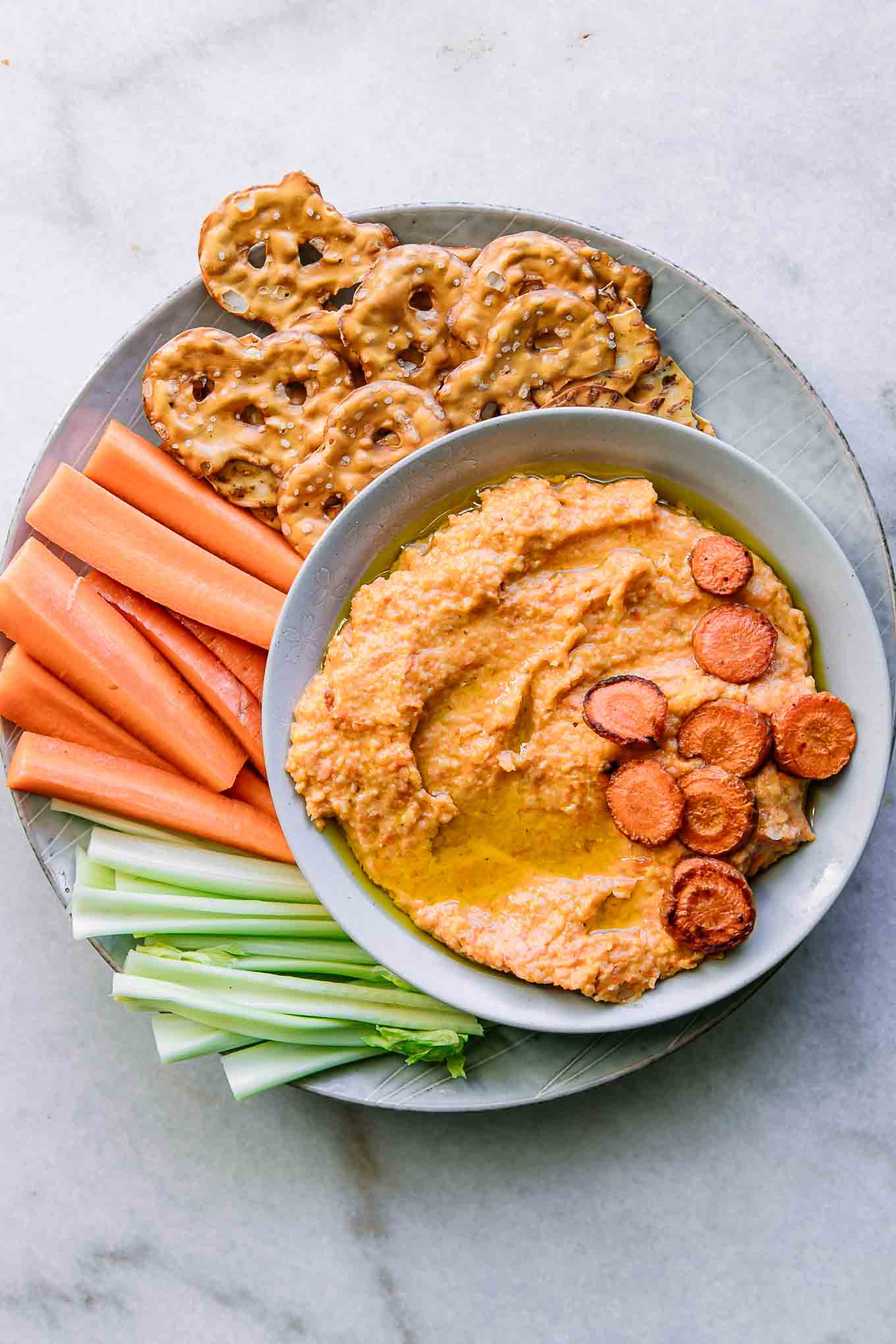 a plate with orange carrot hummus and cut celery, carrots, and pretzels for dipping on a white table
