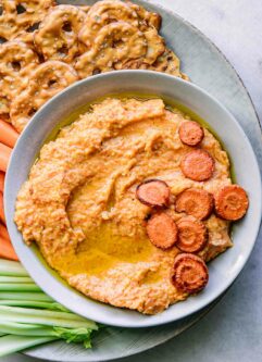 a bowl of orange carrot hummus dip on a plate with celery, carrots, and pretzels