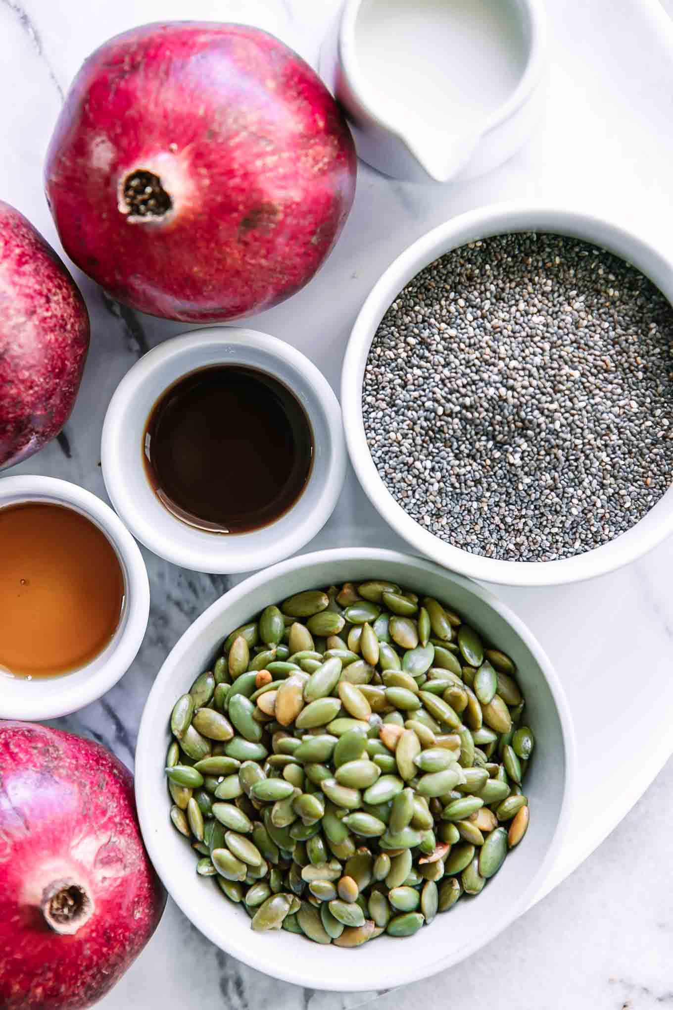 bowls of chia seeds, milk, vanilla, maple syrup, nuts or seeds, and whole pomegranates on a white table for chia seed pudding