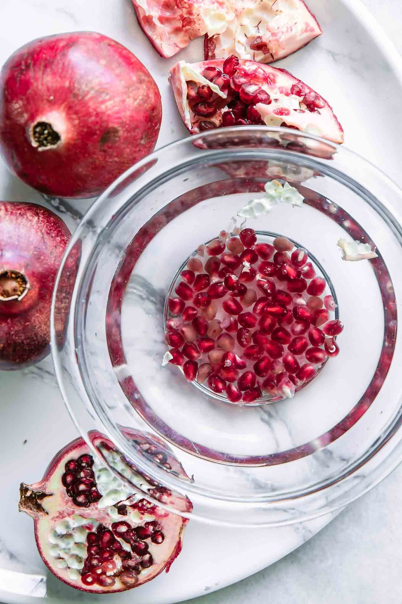 a bowl of water with pomegranate seeds and whole pomegranates on a white table
