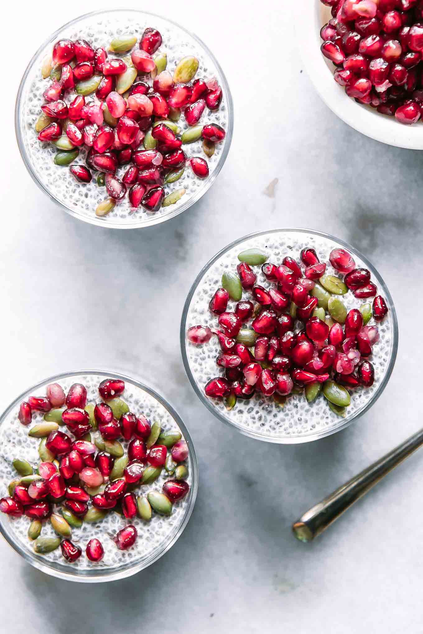 three jars of chia seed pudding with pomegranates and seeds on a white table