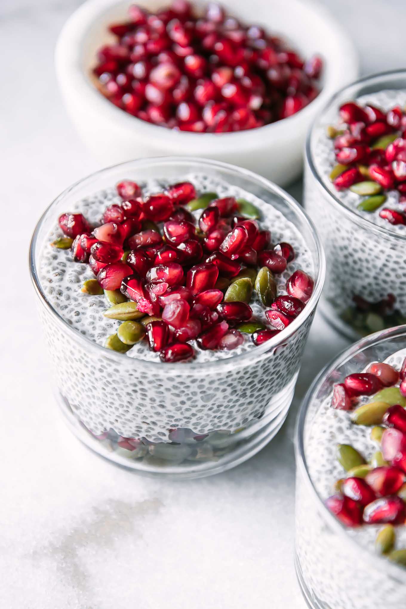 a small glass cup with chia pudding and pomegranates on a white table with a bowl of pomegranate seeds